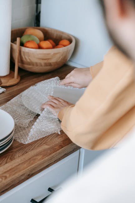 A wooden table filled with packing materials and supplies in a well-lit room, including several stacked rolls of bubble wrap, crumpled sheets of packing paper, a small cardboard box, a black marker, a