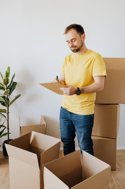 A young woman with curly hair is seated on the floor inside a room during a home relocation process, writing notes on a notepad with a black pen. She is surrounded by packed cardboard boxes, some open